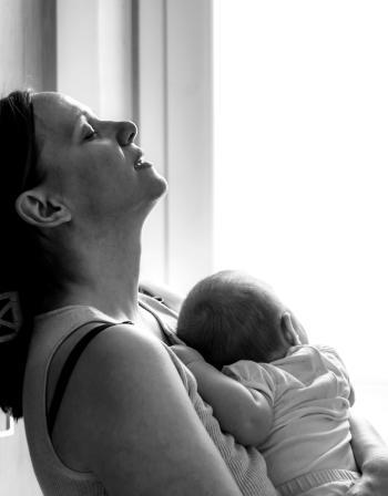 Foto van een vermoeide moeder die tegen de muur aan leunt met haar baby in haar armen, worstelend met vermoeidheid.